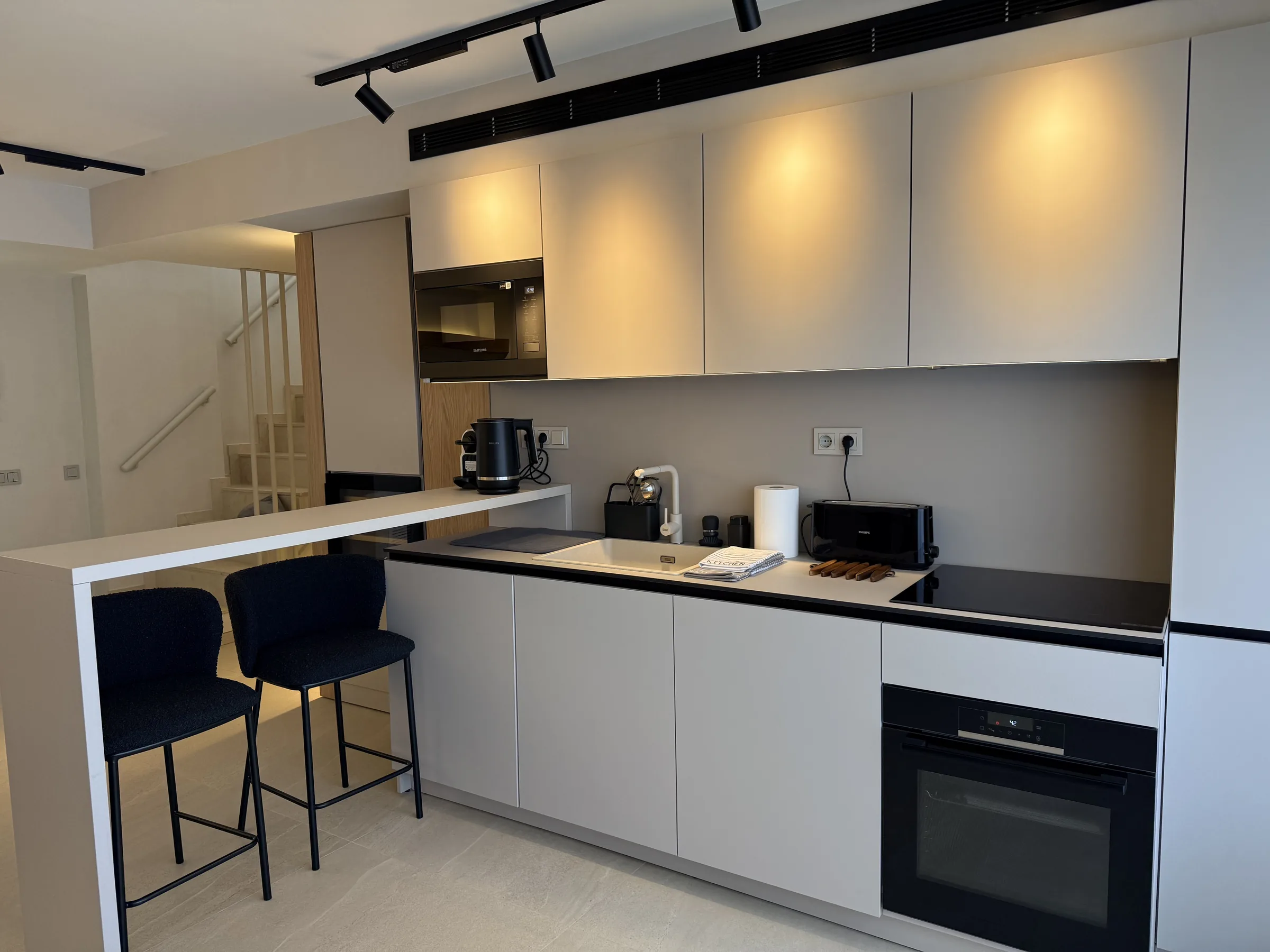 Cocina abierta con barra, muebles blancos y focos de carril en un ático reformado en Platja d'Aro