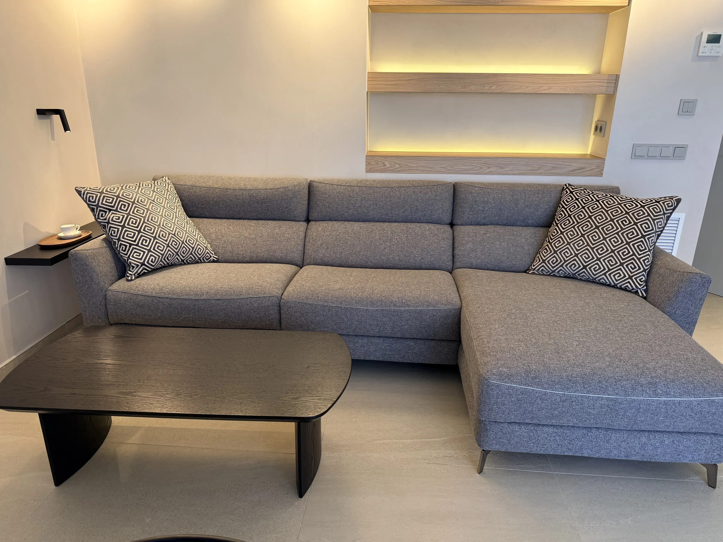 Salón reformado en Platja d'Aro con sofá chaise longue gris, mesa baja y hornacina con estantes de madera iluminados